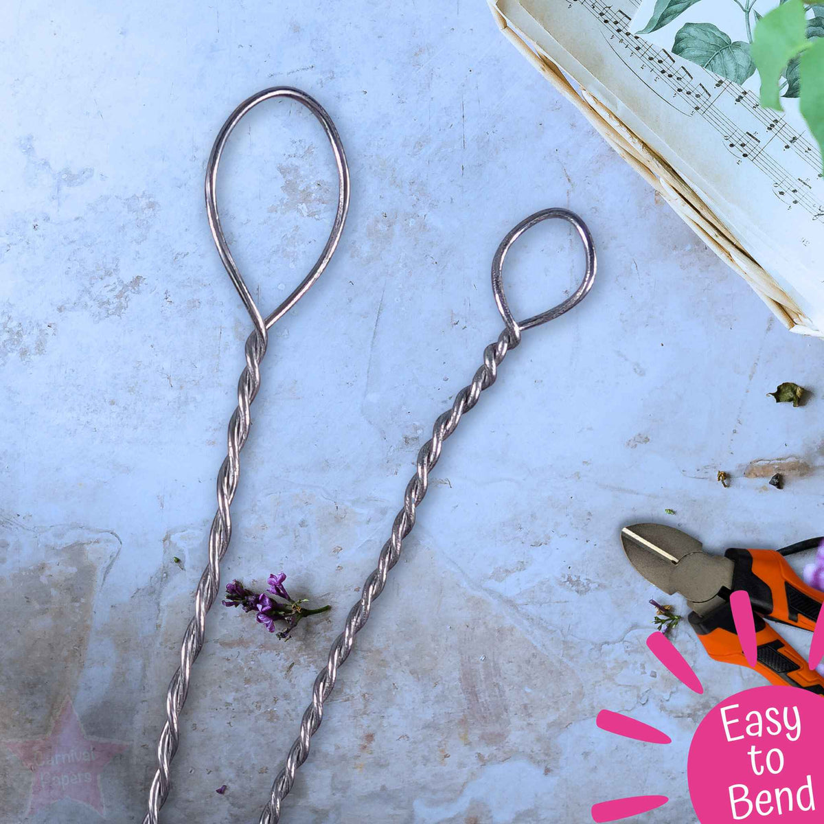 Two twisted metal wire loops with long stems, displayed on a light blue textured surface with small craft props including pliers and a piece of paper.