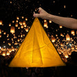 A hand holds up a large, illuminated golden-yellow paper lantern against a night sky filled with thousands of floating lanterns.