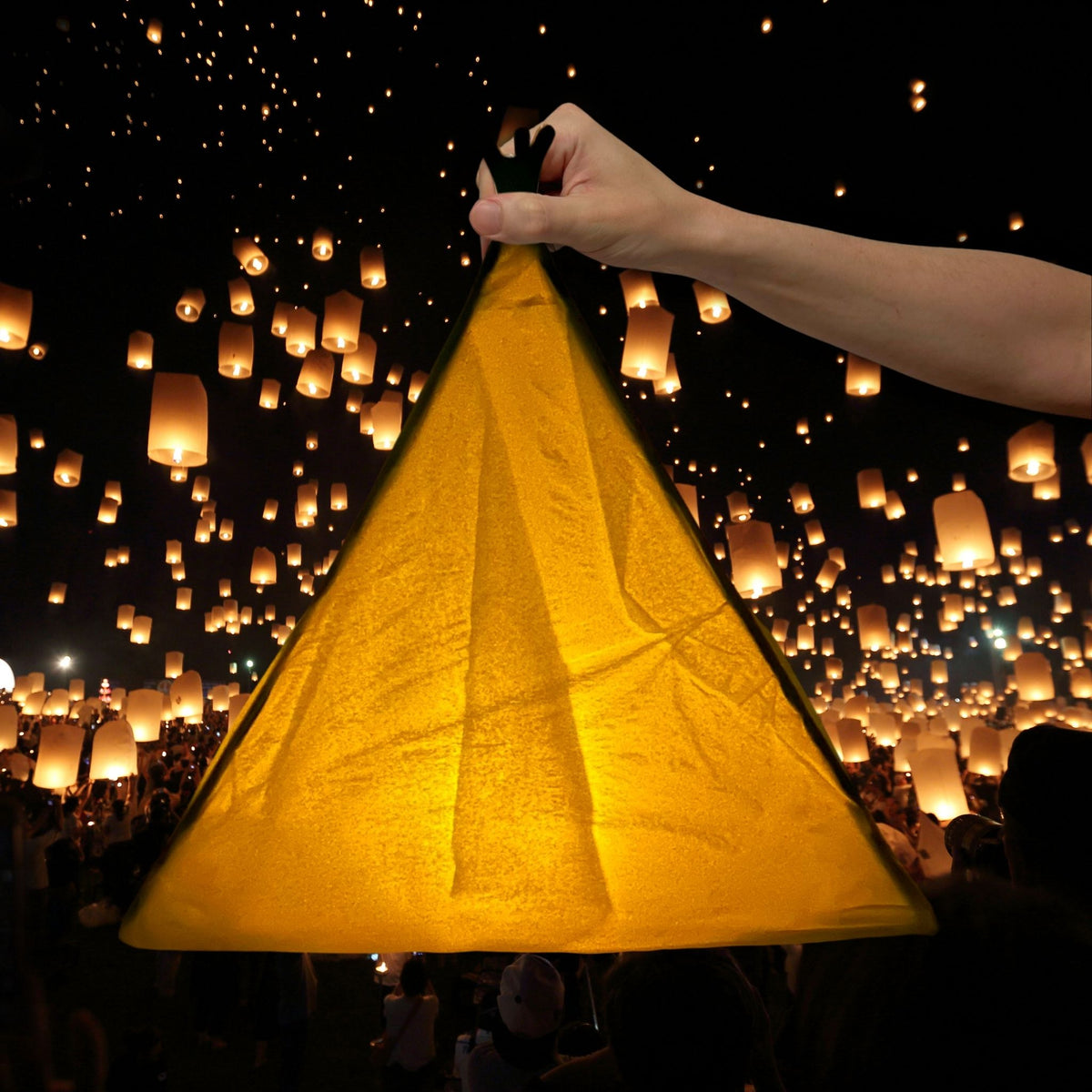 A hand holds up a large, illuminated golden-yellow paper lantern against a night sky filled with thousands of floating lanterns.